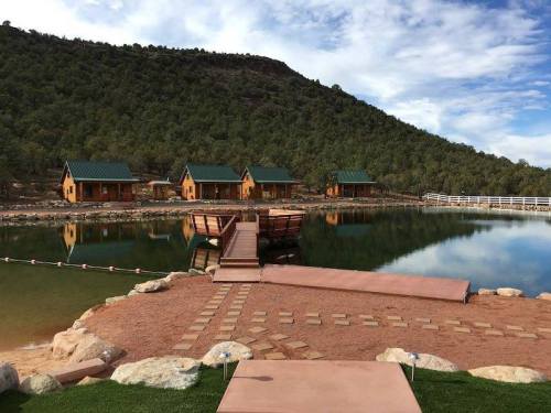 A serene lakeside view featuring wooden cabins, a dock, and a backdrop of green hills under a blue sky.