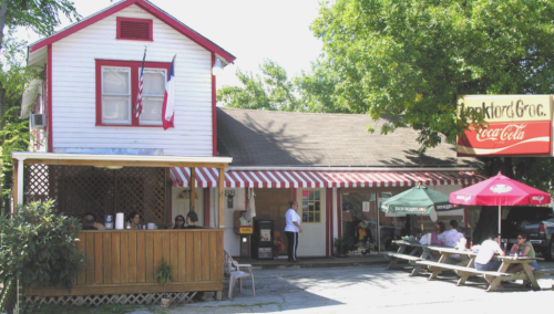 A quaint restaurant with a red and white striped awning, outdoor seating, and a sign for Coca-Cola. Trees surround the building.