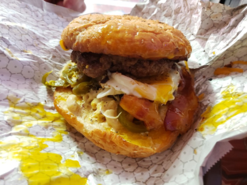 A close-up of a burger with a beef patty, fried egg, jalapeños, and melted cheese, wrapped in paper.