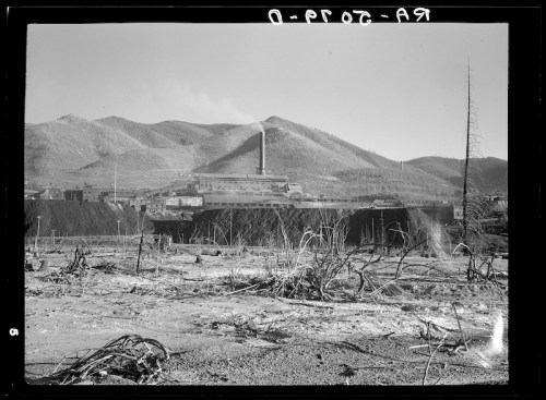A black and white photo of a factory near barren land and mountains, with smoke rising from its chimney.