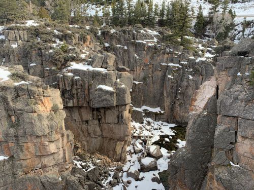 Rocky canyon with snow patches and a small pool of water at the bottom, surrounded by trees and rugged terrain.