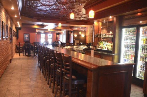 A cozy bar interior featuring a long wooden counter, high stools, and a well-stocked drink fridge against a brick wall.
