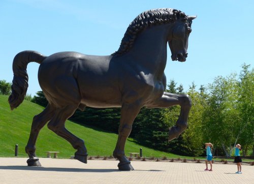 A large bronze horse statue towers over two people standing nearby in a park setting.