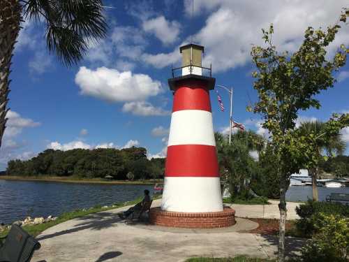 A red and white striped lighthouse by a calm waterway, surrounded by palm trees and blue skies with fluffy clouds.