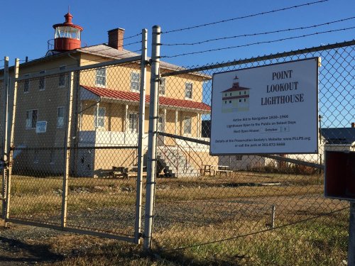 A fenced lighthouse with a red roof and a sign about visiting hours, set against a clear blue sky.