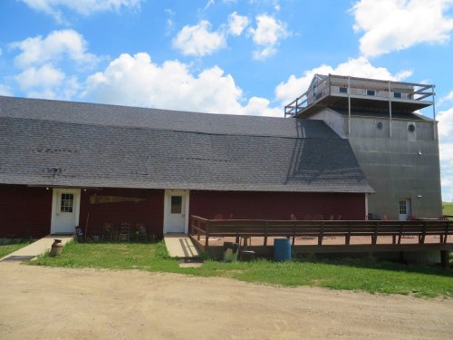 A red barn with a gray grain silo beside it, under a blue sky with fluffy clouds. A wooden deck is in front of the barn.