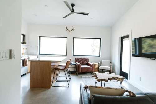Modern living room with a kitchen area, featuring a wooden island, stylish chairs, and large windows.