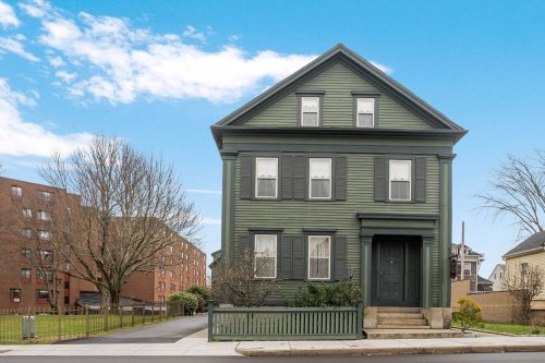 A green, two-story house with black shutters and a front porch, set against a clear blue sky.