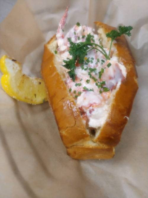 A lobster roll on a toasted bun, garnished with parsley and chives, served with a lemon wedge on the side.