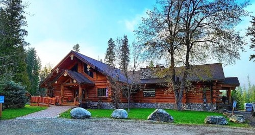 A rustic log cabin surrounded by trees, featuring a stone foundation and a welcoming entrance with a wooden deck.