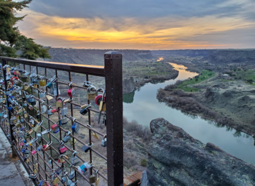 A scenic view of a river winding through hills at sunset, with a fence adorned with colorful padlocks in the foreground.