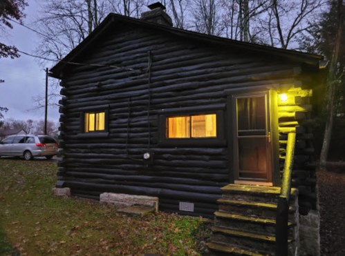 A rustic log cabin with warm lights glowing from the windows, surrounded by trees at dusk.