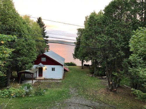 A cozy red and white cabin surrounded by trees, with a serene lake and sunset in the background.