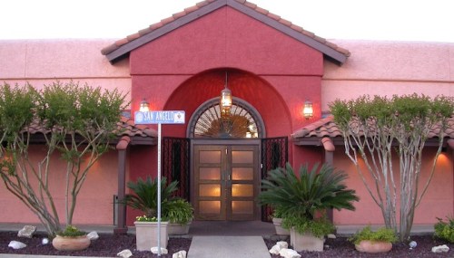 Entrance of a colorful building with a red facade, decorative lanterns, and lush greenery. A street sign reads "San Angel."