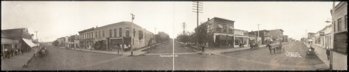 Historic panoramic view of a street scene, featuring buildings, storefronts, and early automobiles.