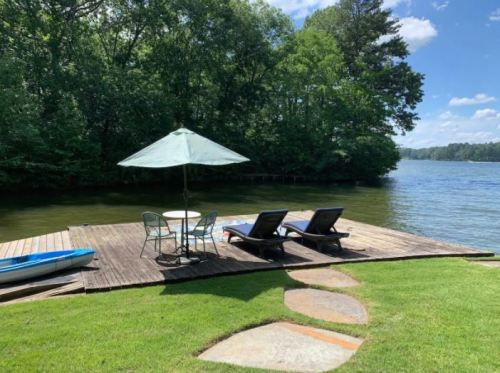 A serene lakeside deck with two lounge chairs, a table under an umbrella, and a kayak nearby, surrounded by greenery.