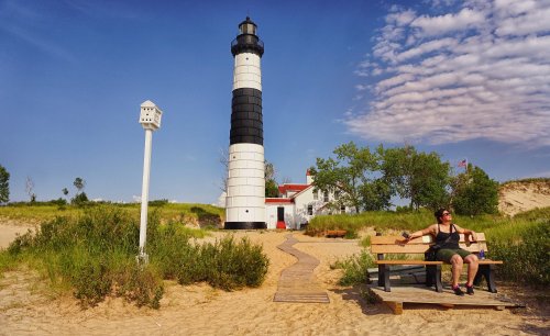 A woman sits on a bench near a black and white lighthouse, surrounded by sandy beach and greenery under a blue sky.