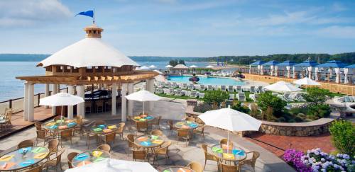 A scenic waterfront resort with a pool, lounge chairs, and a dining area under umbrellas, surrounded by greenery.