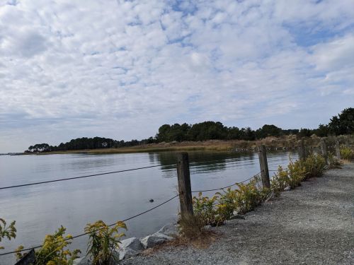 A calm waterway bordered by a gravel path and wooden posts, with greenery and clouds in the sky.