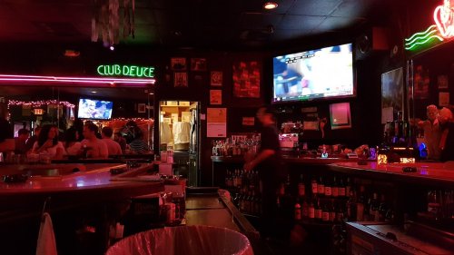 A dimly lit bar with neon signs, a TV showing a game, and a bartender serving drinks.