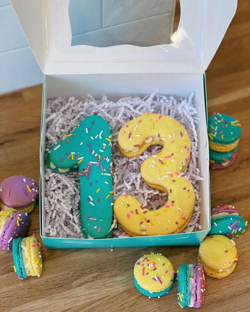 A box containing colorful number cookies "1" and "3" with sprinkles, surrounded by vibrant macarons on a wooden surface.