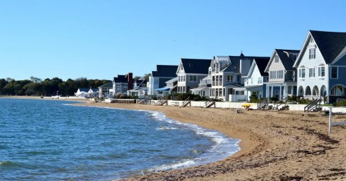 A sandy beach lined with charming houses under a clear blue sky. Gentle waves lap at the shore.