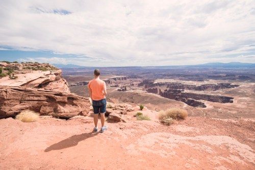 A person stands on a rocky ledge, gazing out over a vast canyon landscape under a cloudy sky.