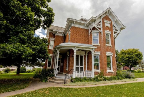 A historic brick house with ornate white trim, surrounded by green trees and a grassy lawn under a cloudy sky.
