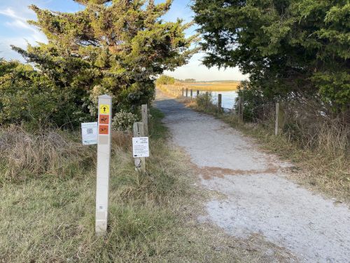 A gravel path leads through greenery towards a body of water, marked by informational signs on either side.