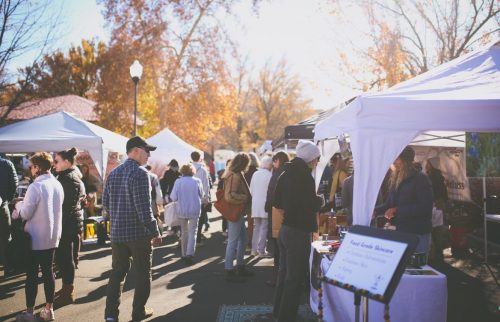 A bustling outdoor market with people browsing tents under sunny autumn skies and colorful trees.