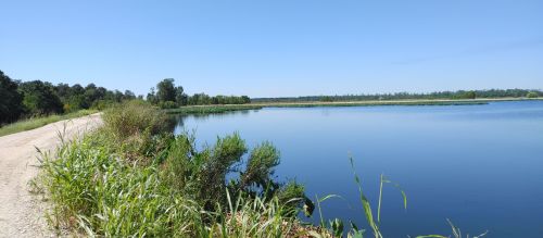 A serene lake surrounded by lush greenery under a clear blue sky, with a gravel path along the water's edge.