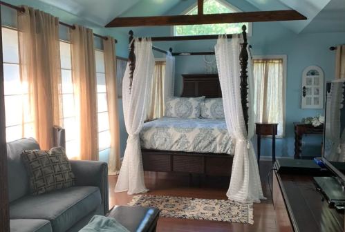Cozy bedroom featuring a four-poster bed with drapes, a gray sofa, and large windows with light curtains.