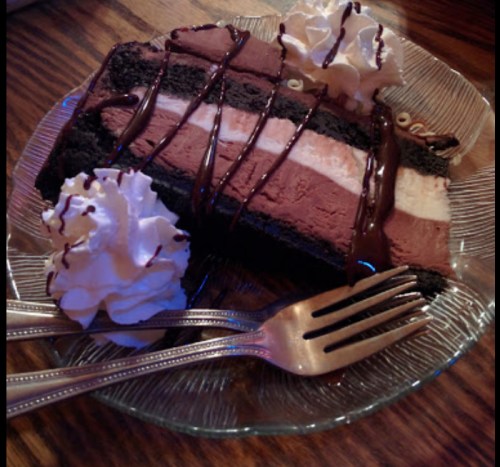 A slice of chocolate mousse cake topped with whipped cream and chocolate drizzle, served on a glass plate.