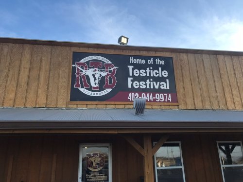 Sign on a wooden building reading "Home of the Testicle Festival" with a logo for R & B Steakhouse.