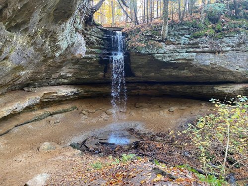A serene waterfall cascades over rocky cliffs into a sandy basin, surrounded by autumn foliage and trees.