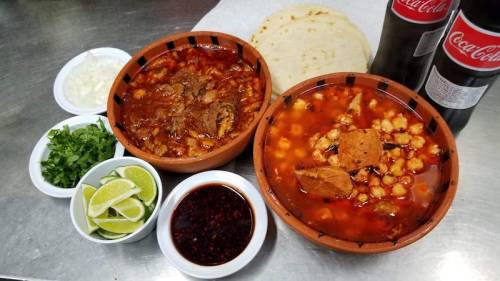 Two bowls of hearty soup with meat, surrounded by lime wedges, chopped onions, cilantro, and tortillas, with Coca-Cola bottles.