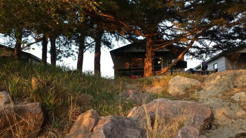 A cabin nestled among trees, with rocky terrain in the foreground, illuminated by warm sunset light.