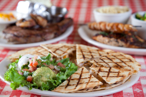 A plate of grilled quesadillas with salad, guacamole, and pico de gallo, with other dishes in the background.