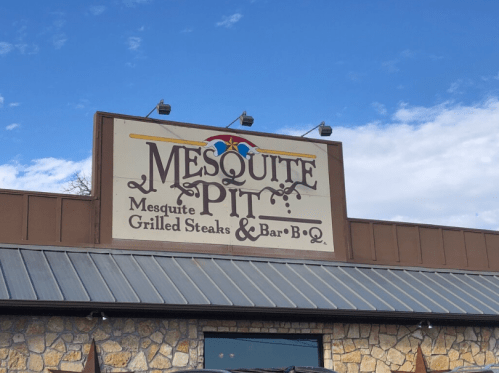 Sign for "Mesquite Pit" featuring grilled steaks and BBQ, set against a blue sky with scattered clouds.