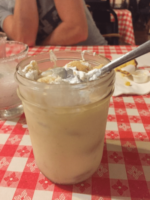 A jar filled with a creamy dessert topped with whipped cream and a spoon, set on a red and white checkered tablecloth.