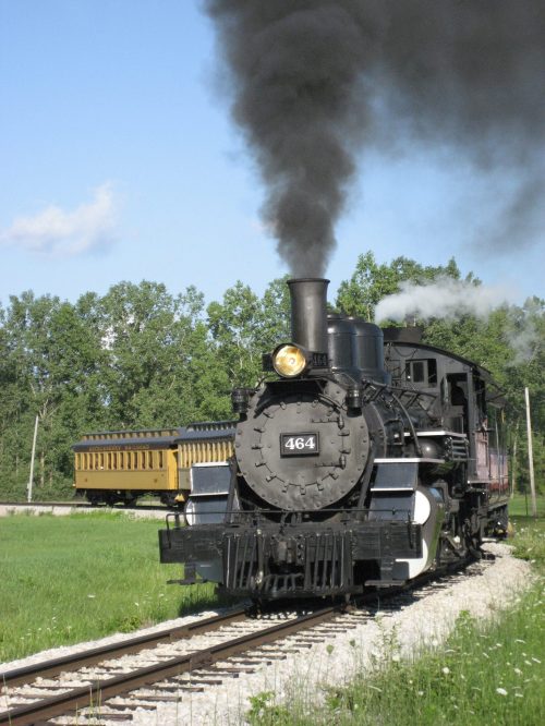 A vintage steam locomotive emits black smoke while pulling a yellow passenger car along a green landscape.