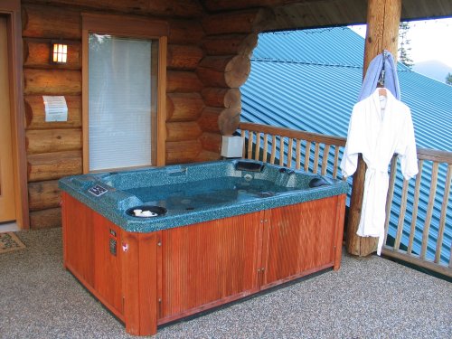 A wooden hot tub on a porch with a bathrobe hanging nearby, surrounded by log cabin walls and a blue roof.
