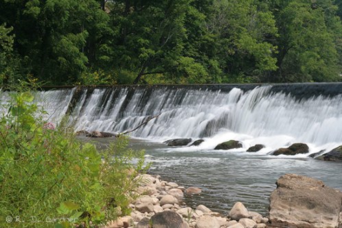 A serene waterfall cascades over rocks, surrounded by lush greenery and a rocky riverbank.