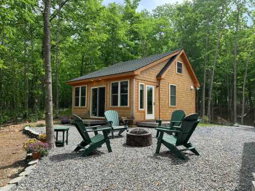 A cozy cabin surrounded by trees, featuring a fire pit and green chairs on a gravel patio.