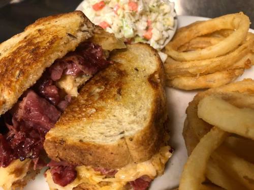 A close-up of a grilled sandwich filled with meat and cheese, served with coleslaw and curly fries on the side.