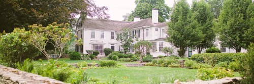 A large white house surrounded by lush greenery and well-maintained gardens on a sunny day.