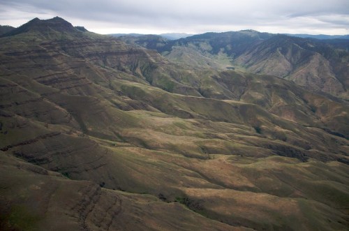 Aerial view of rugged, layered mountains with green and brown slopes under a cloudy sky.