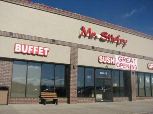 Exterior of Mr. Stirfry restaurant featuring signs for a buffet and sushi, with a bench in front and clear blue sky.