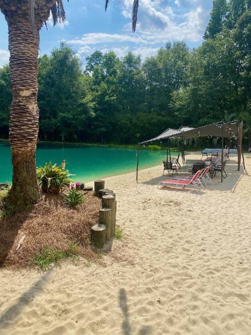 A sandy beach area by a turquoise lake, with palm trees, lounge chairs, and a shaded seating area.