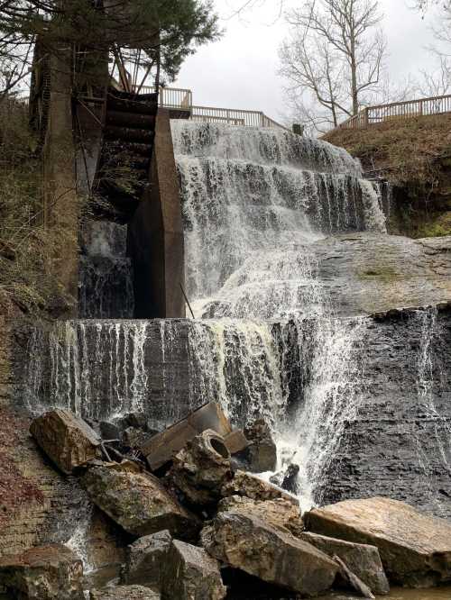 A cascading waterfall flows over rocks, with a structure visible above and trees in the background on a cloudy day.